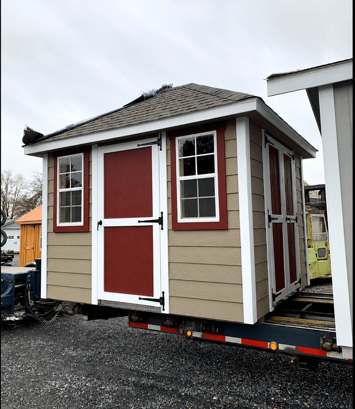 Finished shed with tan lap siding and red doors loaded on a flatbed trailer, ready for delivery
