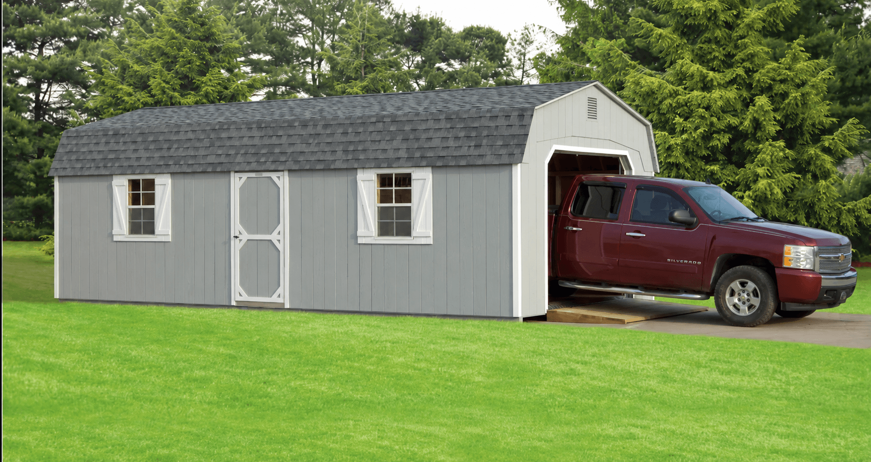 Gray gambrel-roof Amish-built garage with the overhead door open and a pickup truck driving in up a wooden ramp